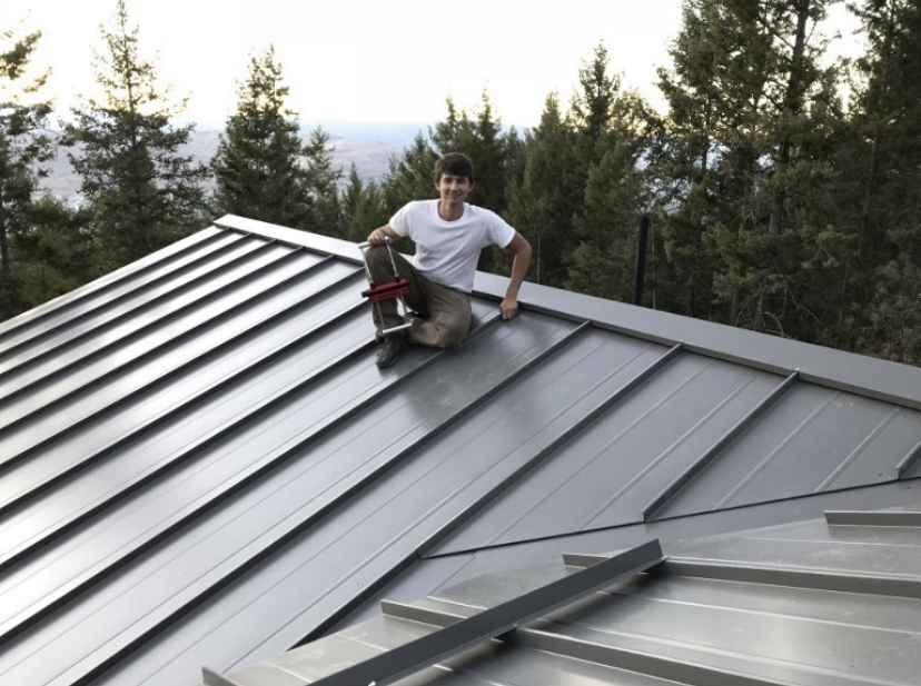 Jared Hanson on a standing seam metal roof he installed, with forest behind